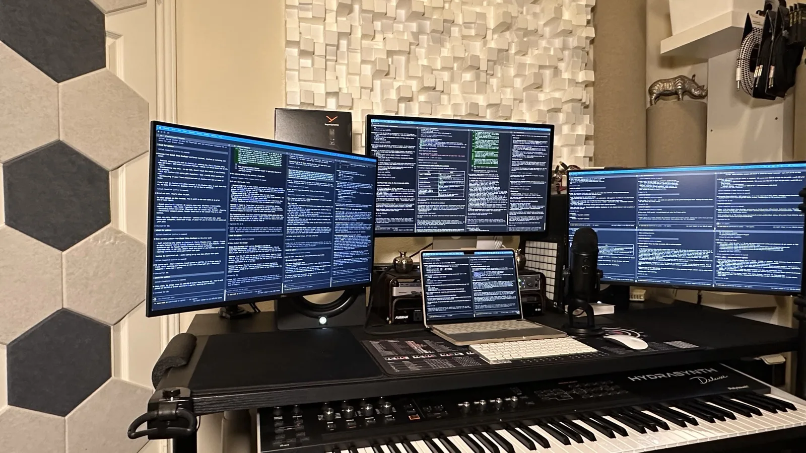 Four-display engineering studio. A laptop is centered on the desk under the middle monitor, flanked by an ultrawide on the left, a wide on the right, and the center display above. All four screens show terminal windows with dense text. A condenser microphone stands to the right of the laptop. The room has acoustic-tile walls.